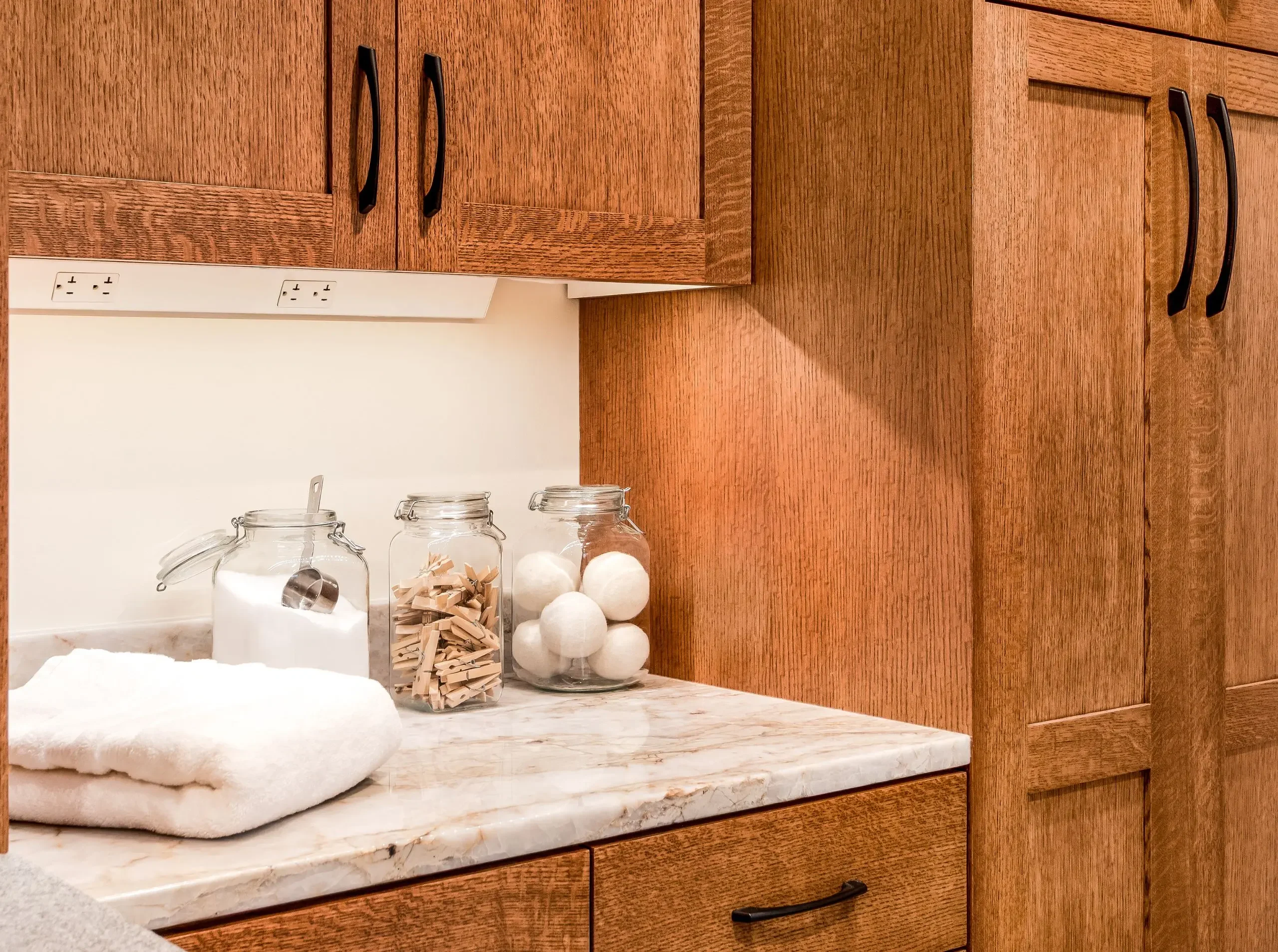 Close-up of a laundry room countertop with glass jars of detergent, clothespins, and dryer balls, set beneath warm wood cabinetry with under-cabinet outlets.