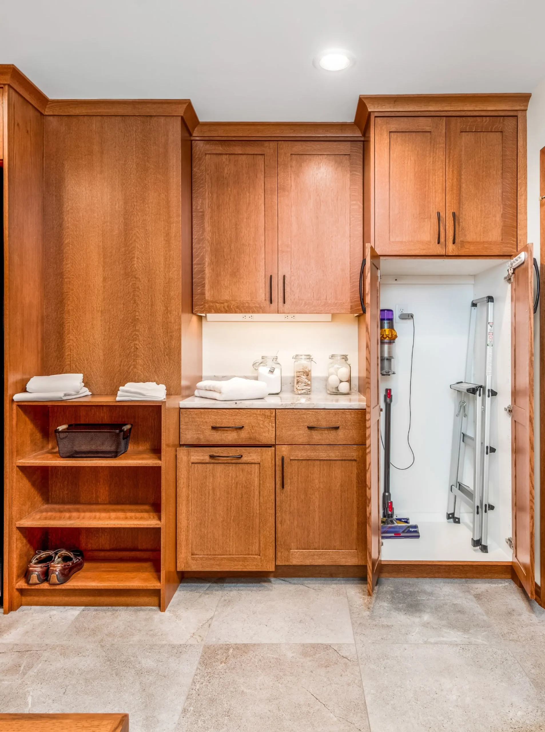 Full view of a custom laundry room with warm wood cabinetry, open shelving, a marble-look countertop, and a tall cabinet opened to reveal a charging station for a vacuum and storage for cleaning tools.